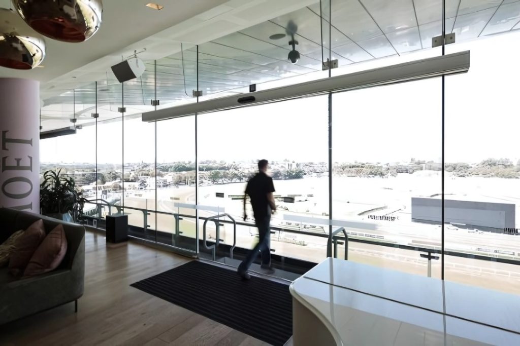 Person walking through full-height glass automatic sliding doors overlooking a racetrack.